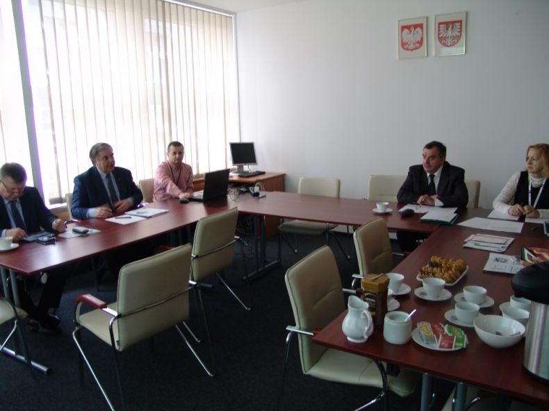 Grupa osób siedzących przy trzech stołach w kształcie litery u. Nad środokowym stołem zawieszone godło Polski i Małopolski.