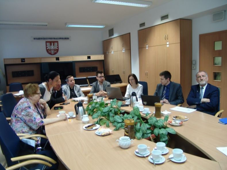 Grupa osób siedzących przy owalnym stole. W tle na ścianie herb województwa małopolskiego.