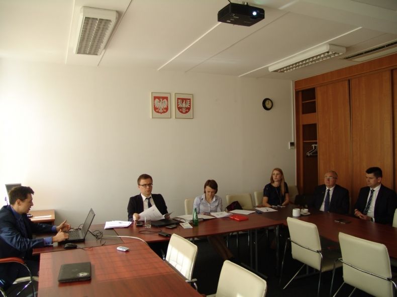 Grupa osób siedzących przy stole w kształcie litery u. Na ścianie nad stołem godło Polski, godło Małopolski i zegar.