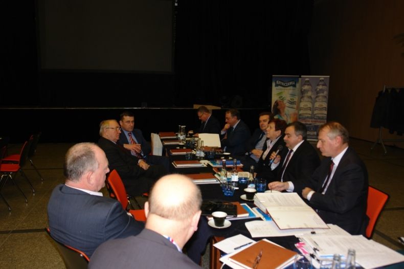Grupa osób siedzi po dwóch stronach stołu. Po prawej stronie radny Jan Piczura z mikrofonem.
