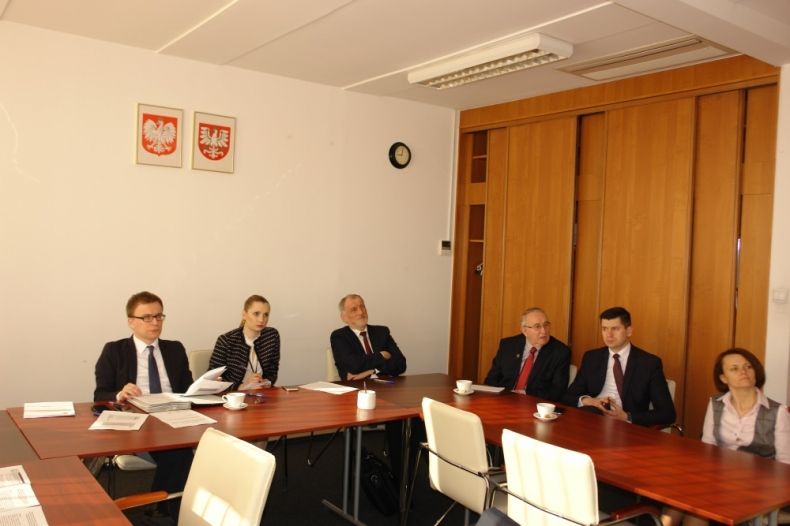 Grupa osób siedzących przy trzech stołach ustawionych w literę U. Na ścianie godła Małopolski i Polski.