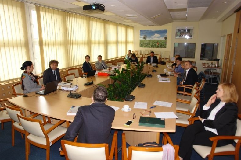 Grupa osób siedzących przy owalnym stole. W tle rząd okien.