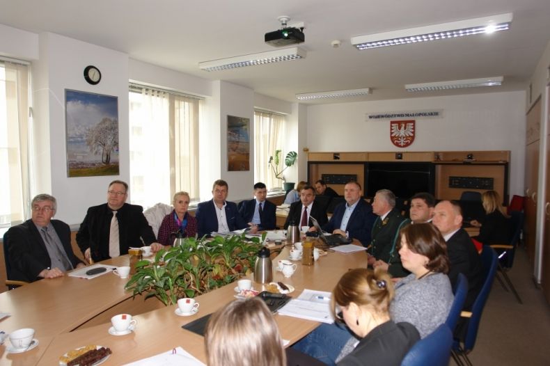 Grupa osób siedzących przy owalnym stole. W tle na ścianie godło województwa.