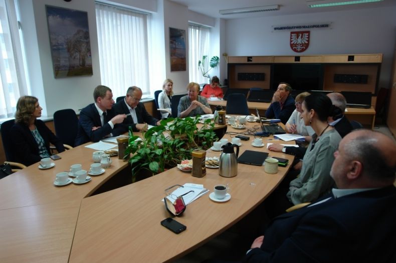 Grupa osób siedzących przy owalnym stole. W tle na ścianie godło województwa.