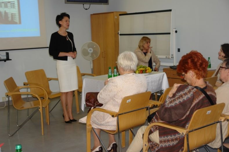 Przewodnicząca Urszula Nowogórska stoi przed stołem i przemawia do mikrofonu. Po drugiej stronie siedzi grupa kobiet.
