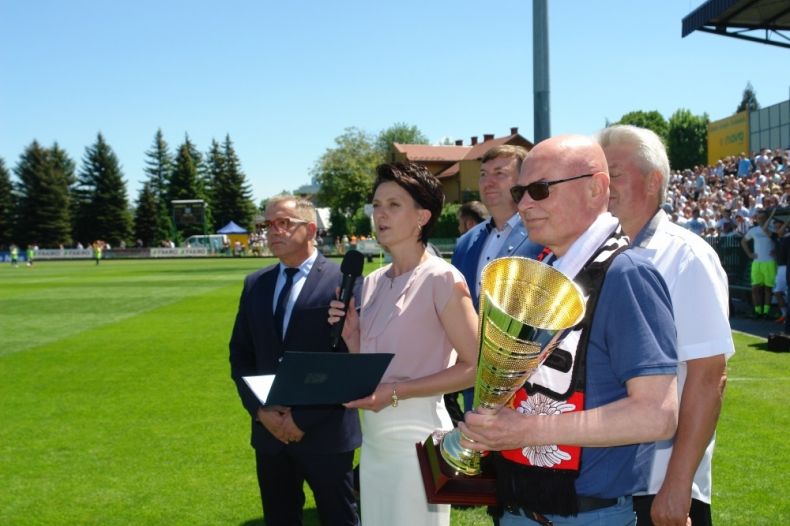 Grupa osób stojących na murawie stadionu. W środku przewodnicząca Urszula Nowogórska trzymająca w ręce granatową teczkę.