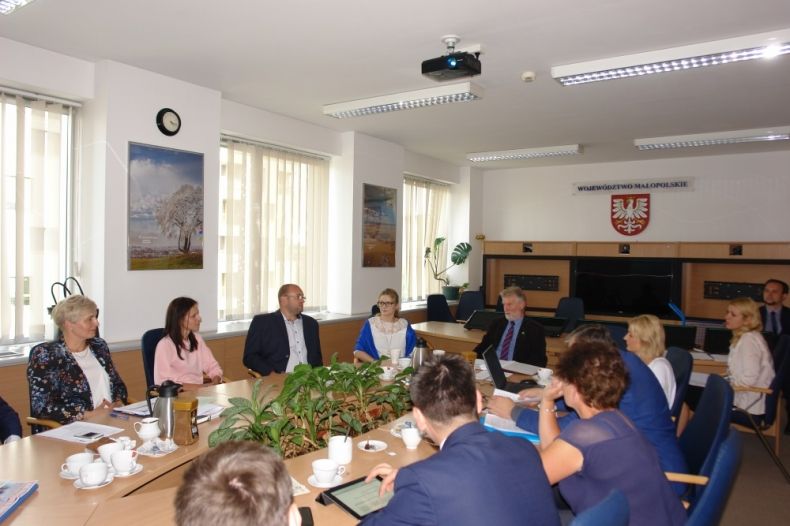 Grupa osób siedzących przy owalnym stole. Na ścianie w tle herb Małopolski.