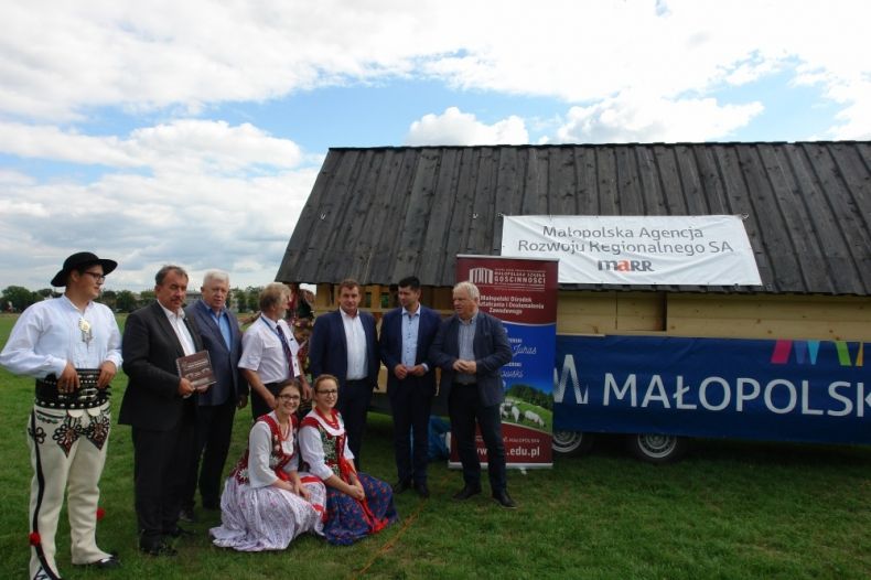 Grupa osób stoi przed drewnianą chatą z napisem Małopolska. Wśród nich osoby w strojach góralskich.