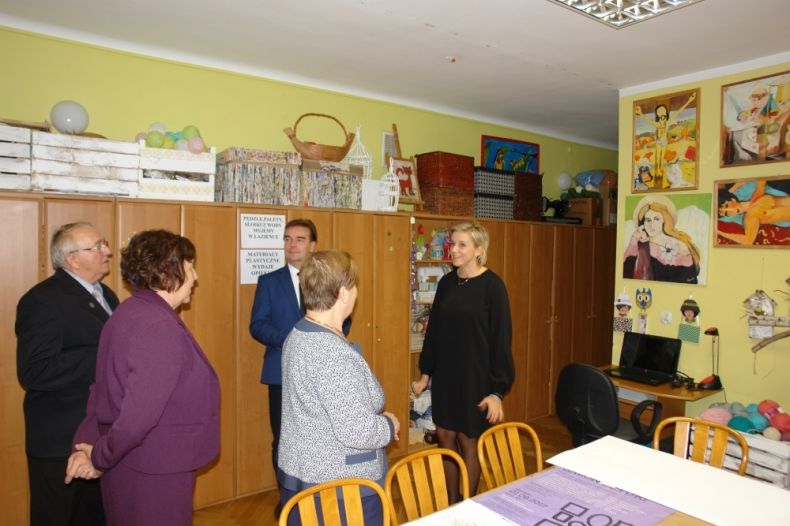 Grupa osób stoi przed ścianą, na której powieszone są prace plastyczne.