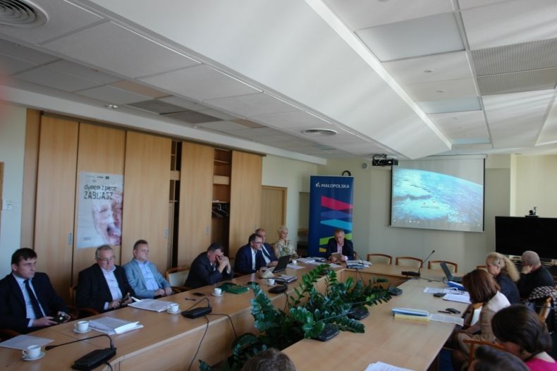 Grupa osób siedzących przy stole. W tle na ścianie wyświetlana jest prezentacja.