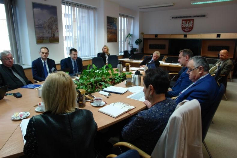 Grupa osób siedzących przy owalnym stole. W tle na ścianie herb województwa.