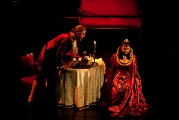Na zdjęciu scena kobieta i mężczyzna przy stoliku. Ona siedzi, on stoi naprzeciw niej