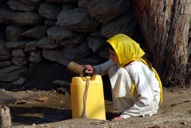 Zdjęcie, dziewczynka kuca nad małym zbiornikiem wodnym. Ma kanister do którego nalewa małym naczyniem wodę. Dziewczynka ma na głowie żółtą chustką