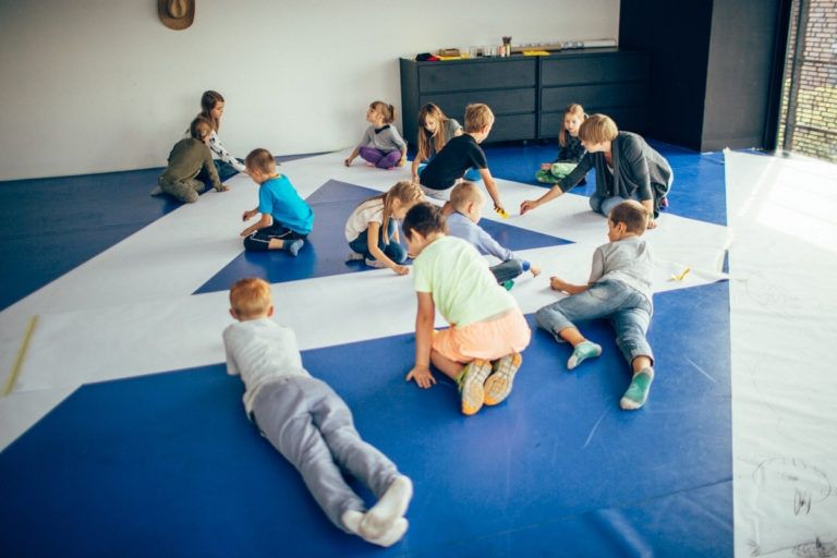 Zdjęcie przedstawia grupę dzieci siedzących i leących na niebieskiej podłodze, wyklejają duży biały trójkąt