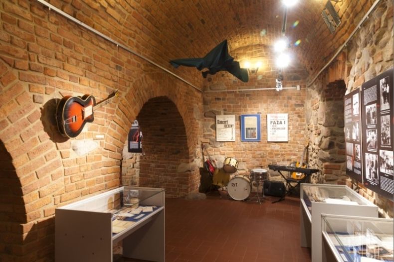 Zdjęcie przedstawia piwnicę, na ścianach ekspozycja wystawy, zdjęcia w gablotach, na ścianie wisi gitara, do sufitu przyczepiona marynarka