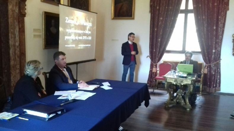 Dwie osoby siedzące przy podłużnym stole i patrzące w prawo na ekran, na którym jest wyświetlana prezentacja. Przed ekranem stoi kolejna osoba.