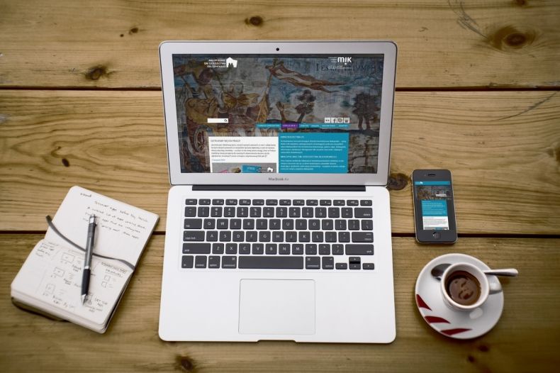 Zdjęcie: Na drewnianym stole laptop, po lewej stronie otatnik z długopisem, po prawej powyżej telefon, poniżej filiżanka z kawą