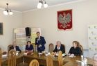 Grupa osób siedzących przy stole. Na ścianie za nimi godło Polski.