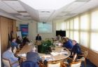 Grupa osób siedzacych przy owalnym stole. W tle na ekranie wyświetlana jest prezentacja.