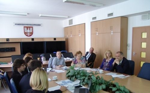 Grupa osób siedzących przy owalnym stole. W tle na ścianie godło Małopolski.