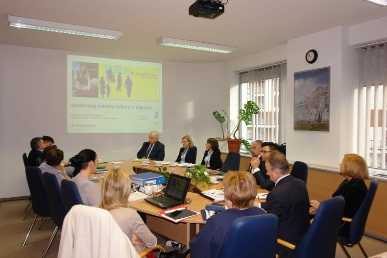 Grupa oósb siedzących przy owalnym stole. W tle na ekranie wyświetlana jest prezentacja.