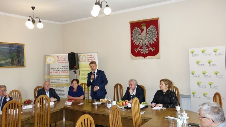 Grupa osób siedzących przy stole. Na ścianie za nimi godło Polski.