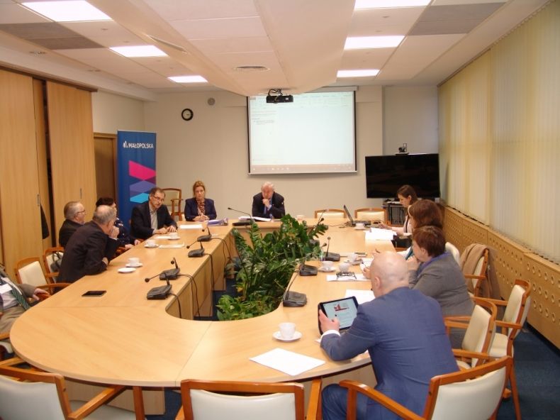 Grupa osób siedzących przy owalnym stole. W tle wyświetlana jest prezentacja.
