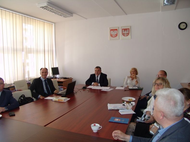 Grupa osób siedzących przy kwadratowym stole. Na ścianie wiszą herby Małopolski i Polski.