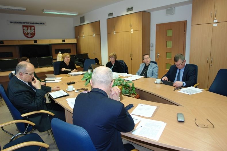 Grupa osób siedzących przy stole. W tle na ścianie wisi godło Małopolski.
