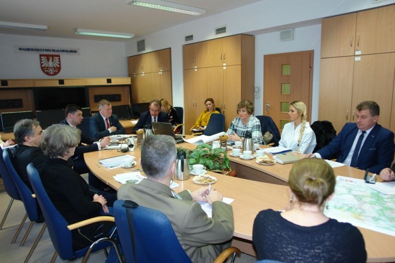 Grupa osób siedzących przy owalnym stole. W tle na ścianie godło Małopolski.