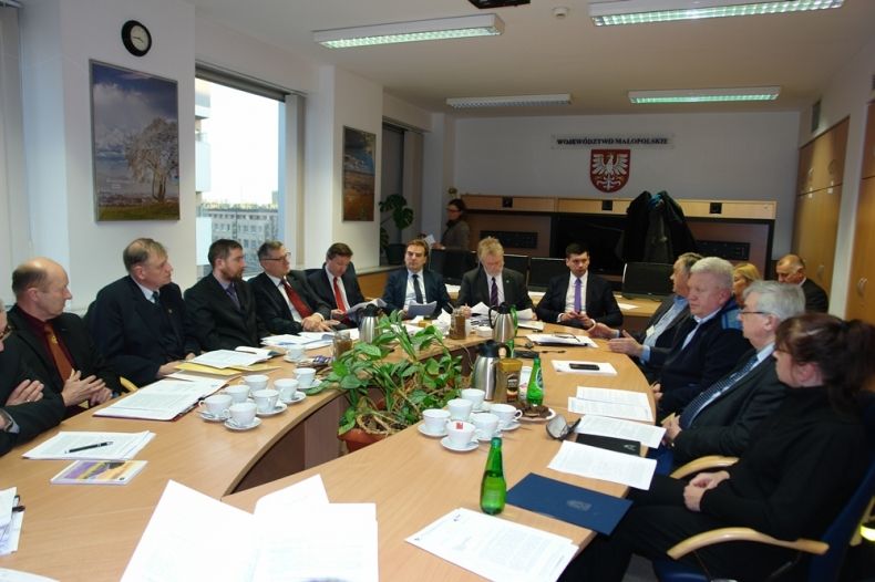 Grupa osób siedzących przy owalnym stole. W tle na ścianie godło Małopolski.