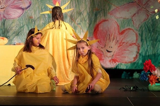 Zdjęcie przedstawia zespół teatralny Stonoga; dwie dziewczynki w żółtych strojach siedzą na podłodze, trzecia stoi odwrócona tyłem. 