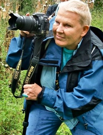 Na zdjęciu przedstawiona jest podróżniczka Elżbieta Dzikowska, podróżniczka. Ubrana jest w niebieską kurtkę i trzyma w dłoni aparat fotograficzny.