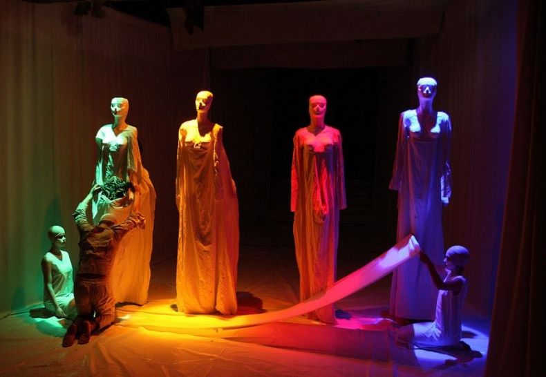 Zdjęcie przedstawia 4 postaci każda ubrana w inny kolor. Postaci stoją na scenie teatru. Obok postaci na podłodze siedzą dwie kobiety ubrane na biało. Przed jedną z postaci klęczy człowiek z rękami uniesionymi do góry.