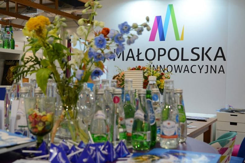 na pierwszym planie wazon z kwiatami i butelki z wodą mineralną z Małopolski, a w tle na białej ścianie logo Małopolska Innowacyjna