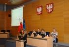 Cztery osoby za stołem prezydialnym. Poniżej jedna osoba przy mównicy. Na ekranie po prawej wyświetlana jest prezentacja, pod nią flagi Małopolski, Polski i Unii Europejskiej. Na ścianie godła Polski i Małopolski.