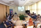 Grupa osób siedzących przy owalnym stole. W tle na ekranie wyświetlana jest prezentacja.