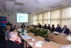 Grupa osób siedzących przy owalnym stole. W tle na ekranie wyświetlana jest prezentacja.