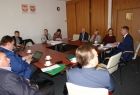 Grupa osób siedzących przy prostokątnym stole. W tle na ścianie godła Małopolski i Polski.