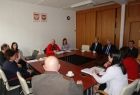 Grupa osób siedzacych przy kwadratowym stole. W tle na ścianie godła Małopolski i Polski.