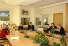 Grupa osób siedzących przy owalnym stole. W tle na ścianie zdjęcie przedstawiające łąkę i niebieskie niebo.