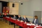Grupa osób siedzących przy podłóżnym stole z bordowym obrusem.