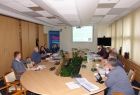 Grupa osób siedzących przy owalnym stole. W tle na ekranie wyświetlana jest prezentacja.