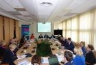 Grupa osób siedzących przy owlanym stole. W tle na ekranie wyświetlana jest prezentacja.