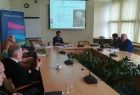 Grupa osób siedzących przy prostokątnym stole. W tle na ekranie wyświetlana jest prezentacja.