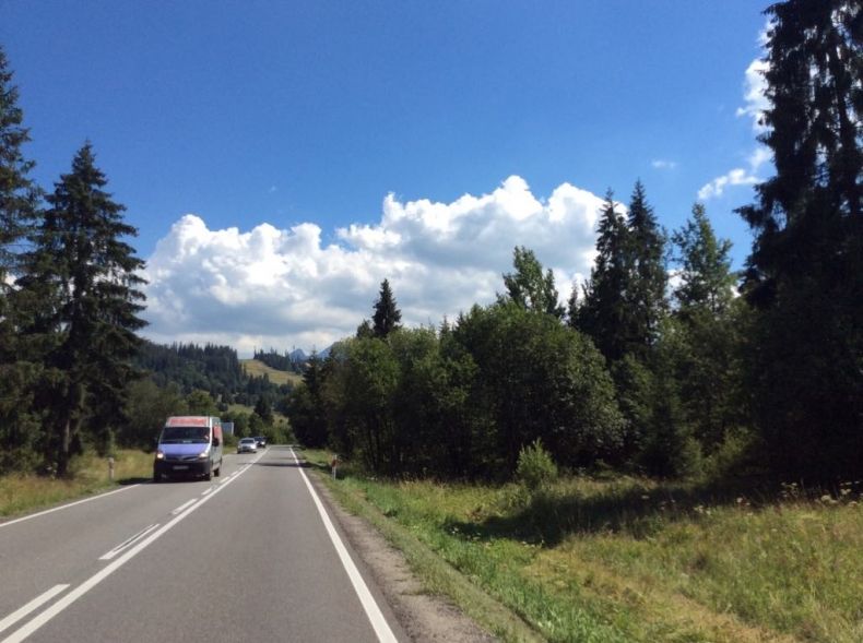 droga asfaltowa dla samochodów, na poboczu rosną drzewa, nad jezdnią jest błękitne niebo, w tle nadjeżdżający bus