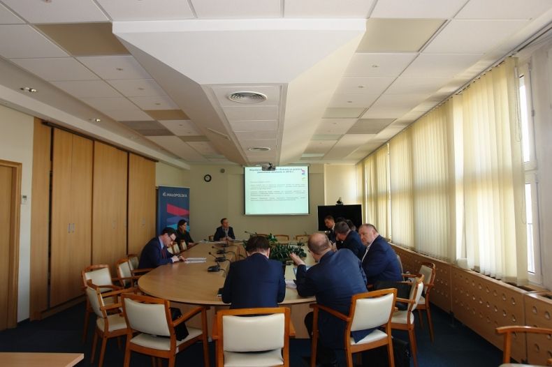 Grupa osób siedzących przy owalnym stole. W tle na ekranie wyświetlana jest prezentacja.
