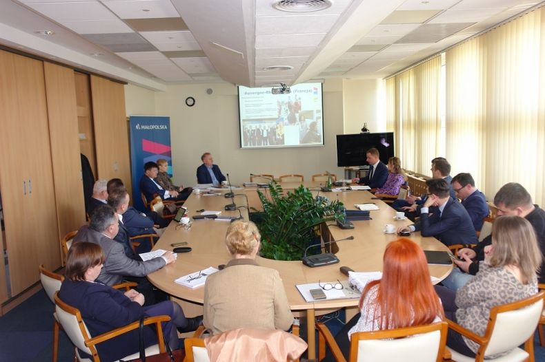 Grupa osób siedzących przy owalnym stole. W tle na ekranie wyświetlana jest prezentacja.