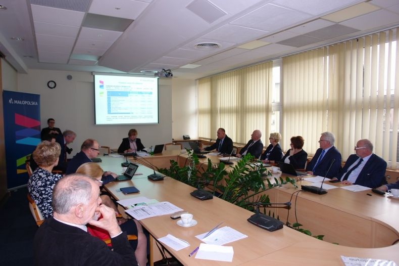 Grupa osób siedzących przy owalnym stole. W tle na ekranie wyświetlana jest prezentacja.