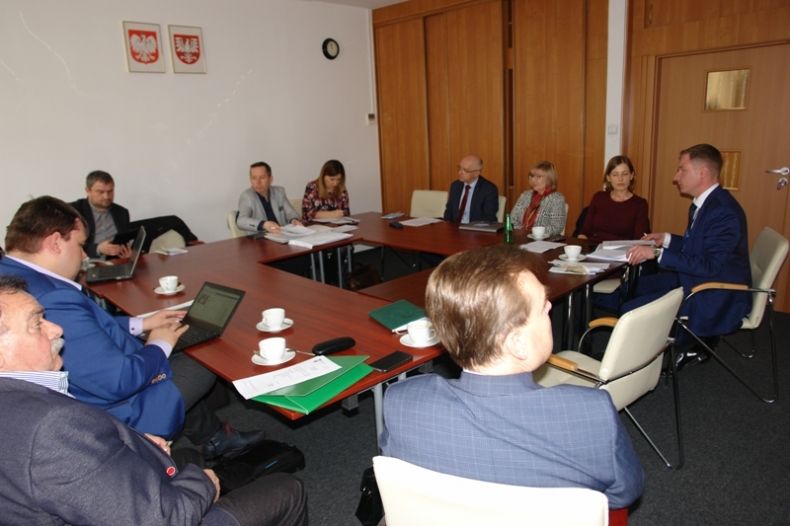 Grupa osób siedzących przy prostokątnym stole. W tle na ścianie godła Małopolski i Polski.
